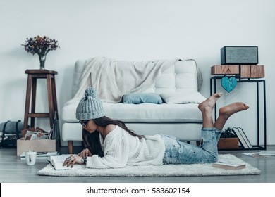 Reading Interesting Story. Beautiful Young Woman In Casual Wear And Knit Hat Reading Book While Lying On The Floor At Home