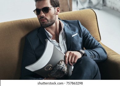 Reading Latest News. Good Looking Young Man In Full Suit Holding A Magazine And Looking Away While Sitting On The Sofa