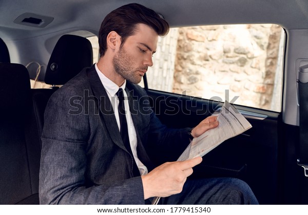 Reading latest news. Handsome\
young man in full suit reading a newspaper while sitting in the\
car.