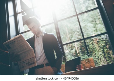 Reading Latest News. Low Angle View Of Confident Young Man Reading Fresh Newspaper While Leaning At The Window Sill In Office Or Cafe