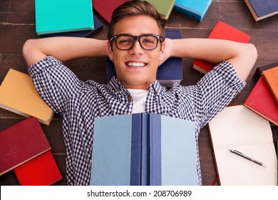 Reading Is My Hobby! Top View Of Happy Young Man Holding Hands Behind Head And Smiling While Lying On The Hardwood Floor With Colorful Books Laying All Around Him 