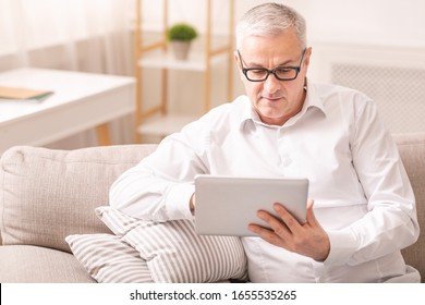 Reading News. Senior man in spectacles using digital tablet sitting on the couch at home, copyspace - Powered by Shutterstock