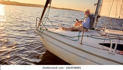 Reading on board. Side view of a relaxed senior man sitting on the side of sailboat or yacht deck floating in the calm blue sea at sunset and using digital tablet or ebook.