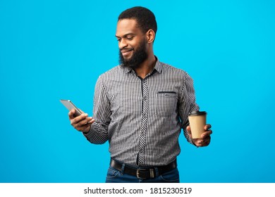 Reading Sms. Smiling Black Business Man Texting On Cell Phone Standing Over Blue Studio Background, Holding Paper Coffee Cup. Bearded African American Guy Chatting Online Using His Gadget, Copy Space