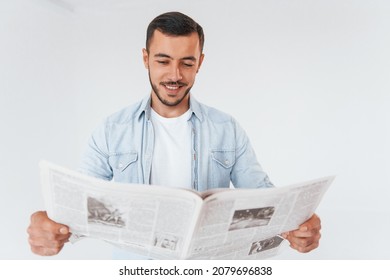 Reads Newspaper. Young Handsome Man Standing Indoors Against White Background.