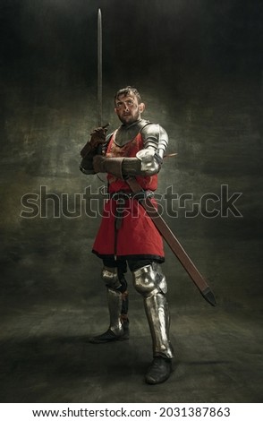Ready for defence. Portrait of one brutal bearded man, medeival warrior or knight with dirty wounded face holding big sword isolated over dark background.