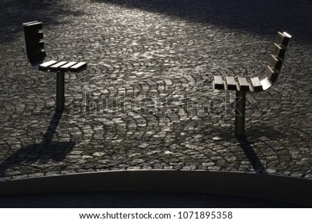 Ready for dialog. Two single chairs installed opposite to each other in a city square as benches. Modern cityscape or urban environment. Backlit black-and-white outdoor photograph featuring pavement.