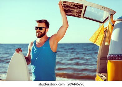 Ready to have some fun. Smiling young man holding skimboard and while opening a trunk door of his retro minivan with sea in the background  - Powered by Shutterstock