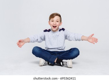 Ready To Hug The World. Happy Boy In Braces Sitting Legs Crossed On Studio Floor. Kid In Casual Laughing, Posing On Camera At White Background, Copy Space