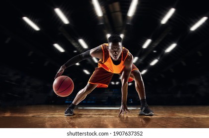 Listo para saltar. Joven jugador de baloncesto afroamericano en acción y movimiento en linternas sobre fondo oscuro de gimnasio. Concepto de deporte, movimiento, energía y estilo de vida dinámico y saludable. El estropajo de Arena