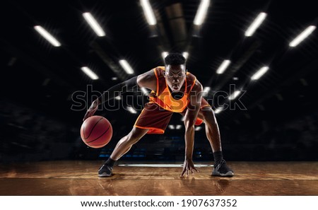 Ready to jump. African-american young basketball player in action and motion in flashlights over dark gym background. Concept of sport, movement, energy and dynamic, healthy lifestyle. Arena's drawned