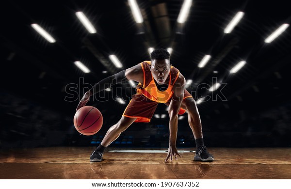Ready to jump. African-american young basketball\
player in action and motion in flashlights over dark gym\
background. Concept of sport, movement, energy and dynamic, healthy\
lifestyle. Arena\'s\
drawned