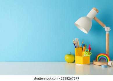 Ready to learn: side view photo of a desk featuring school supplies, pencil holder, pens, ruler, scissors, rainbow plasticine, adhesive tape, lamp, fresh apple against a blue wall with space for text