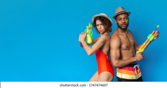 Ready For Pool Battle. Focused african guy and girl in swimwear posing with water guns on blue background, panorama with copy space