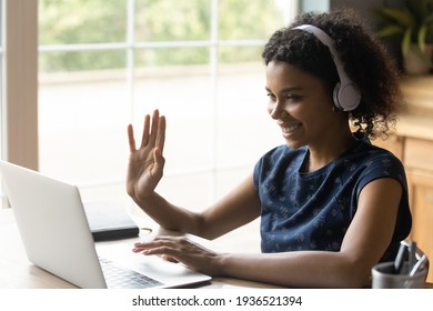 Ready to start. Positive biracial teen female in headset sit by laptop waving hand to camera greet teacher tutor at distant video lesson. Young black woman student learning online via web conference