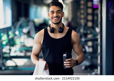 Ready For Training. Portrait Of Happy Young Sporty Arab Guy Posing At Gym Interior, Handsome Middle Eastern Male Athlete Holding Towel And Sport Shaker Bottle And Smiling At Camera, Copy Space