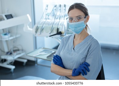 Ready For Treatment. Pretty Young Female Dentist Wearing Safety Goggles, Face Mask And Rubber Gloves While Posing For The Camera And Folding Her Hands Across Her Chest