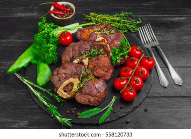 Ready-cooked beef meat Osso buco Veal shank on black stone board. Ingredients for Osso buco thyme, parsley, rosemary, lettuce, pepper, sage. Dark wooden background. - Powered by Shutterstock