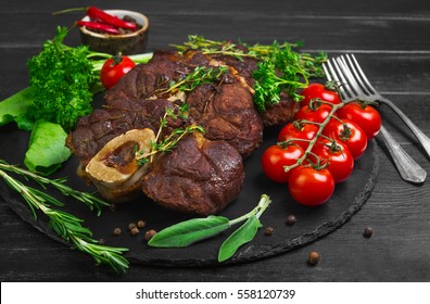 Ready-cooked beef meat Osso buco Veal shank on black stone board. Ingredients for Osso buco thyme, parsley, rosemary, lettuce, pepper, sage. Dark wooden background. - Powered by Shutterstock