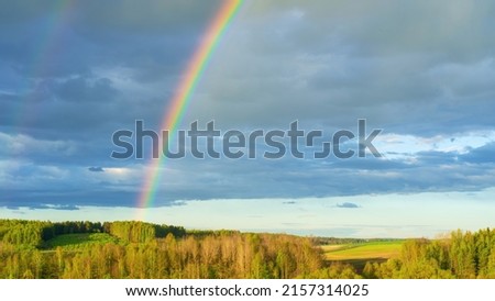 A real double rainbow in a cloudy sky after heavy rain. A rare natural phenomenon in the blue sky. Rainbow over hilly countryside in early spring. The concept of weather forecast and climate change.