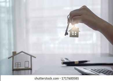 Real Estate Agent Holding House Key On House Shaped Keyring On Table With House Designs Document, Calculator, Model House. Concept Of Investment Property.