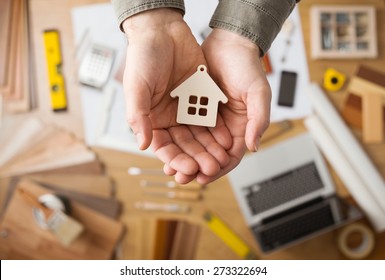 Real Estate Agent Holding A Small House, Desktop With Tools, Wood Swatches And Computer On Background, Top View