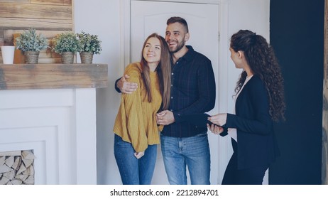 Real estate agent is meeting happy married couple, showing papers and telling them about new apartment. Young people are excited, they are looking around and hugging. - Shutterstock ID 2212894701