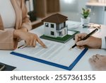 Real estate agent pointing at contract terms, explaining agreement details to client before signing, with house model on desk