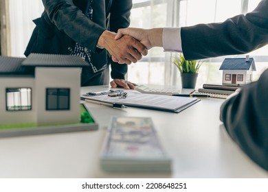 A real estate agent shakes hands with a client after completing a transaction regarding a house contract agreement. Contract documents and a house model on a wooden table. - Shutterstock ID 2208648251