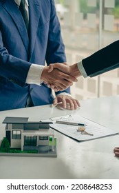A real estate agent shakes hands with a client after completing a transaction regarding a house contract agreement. Contract documents and a house model on a wooden table. - Shutterstock ID 2208648253