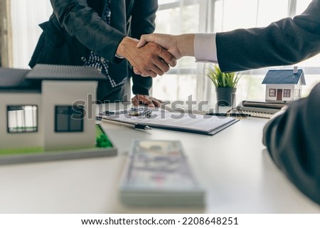 A real estate agent shakes hands with a client after completing a transaction regarding a house contract agreement. Contract documents and a house model on a wooden table.