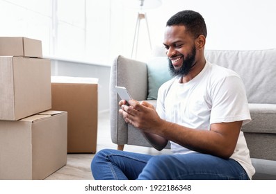 Real Estate App. Happy Black Man Using Phone Browsing Internet Searching New Apartment For Rent Via Mobile Application Sitting Among Packed Moving Boxes At Home. House Purchase And Ownership