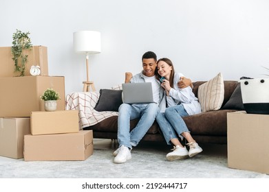 Real Estate Concept. Happy Mixed Race Family Couple Sitting On A Sofa Between Cardboard Boxes In Living Room Of Their New Home, Using Laptop And Credit Card For Online Shopping, Buying Things For Home