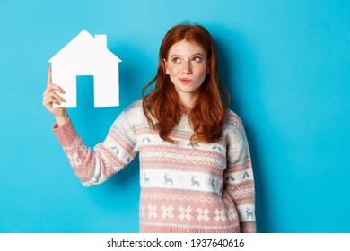 Real estate concept. Image of thoughtful redhead girl showing paper house model and thinking, searching for home or flat, standing against blue background
