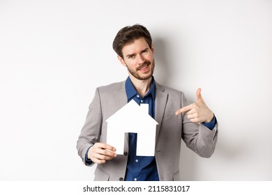 Real Estate And Insurance Concept. Salesman In Grey Suit Showing Paper House Cutout, Selling Property, Smiling At Camera, White Background