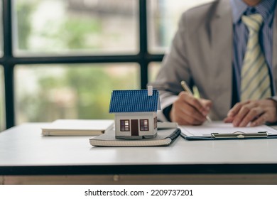 Real Estate Office, Sales Representatives Sitting At A Desk With Contract Documents And A Laptop And A Small House Next To It.