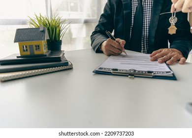 Real Estate Office, Sales Representatives Sitting At A Desk With Contract Documents And A Laptop And A Small House Next To It.