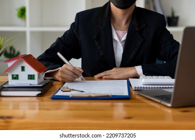 Real Estate Office, Sales Representatives Sitting At A Desk With Contract Documents And A Laptop And A Small House Next To It.