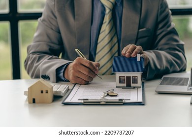 Real Estate Office, Sales Representatives Sitting At A Desk With Contract Documents And A Laptop And A Small House Next To It.