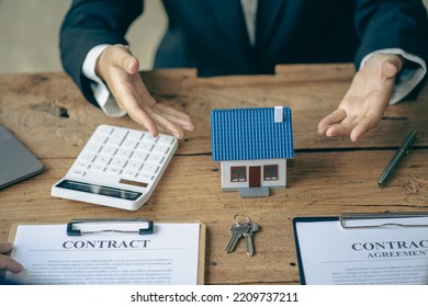 Real Estate Office, Sales Representatives Sitting At A Desk With Contract Documents And A Laptop And A Small House Next To It.