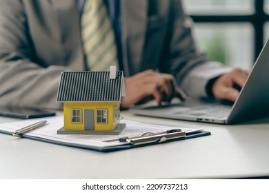 Real Estate Office, Sales Representatives Sitting At A Desk With Contract Documents And A Laptop And A Small House Next To It.