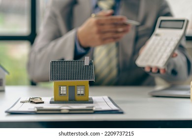 Real Estate Office, Sales Representatives Sitting At A Desk With Contract Documents And A Laptop And A Small House Next To It.