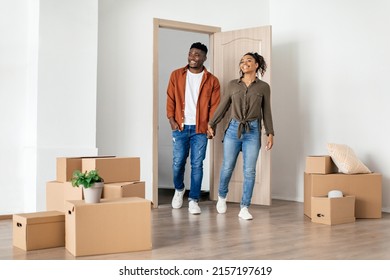 Real Estate Ownership. Happy Black Spouses Holding Hands Entering New Home, Looking At Empty Living Room And Cardboard Boxes Moving Apartment. Family Housing Concept. Full Length