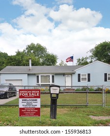 Real Estate For Sale Sign American Dream Great Starter Home Affordable First Time Buyers Welcome Open House Residential Neighborhood USA American Flag Chain Link Fence Blue Sky Clouds