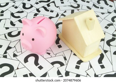 Real Estate And Saving Money Questions Concept. Wooden House Model With Piggy Bank On Question Marks Background.