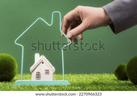 Real estate services. House insurance protection and safety. moving and relocation. Hand with small house within bigger house frame in green residential area. Loan for new home. Finding new property