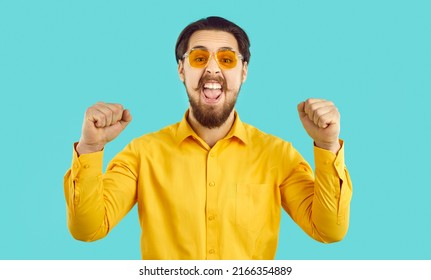 Real Happiness. Happy Stylish Funny Funky Crazy Hipster Guy Shouting With Joy On Light Blue Background. Excited Bearded Man In Orange Shirt And Sunglasses Joyfully Shouts At Camera Clenching Fists.