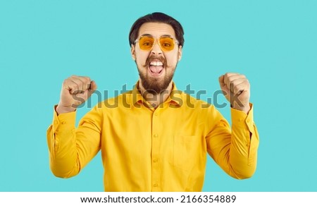Real happiness. Happy stylish funny funky crazy hipster guy shouting with joy on light blue background. Excited bearded man in orange shirt and sunglasses joyfully shouts at camera clenching fists.