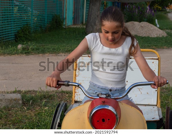 Real\
life. A beautiful girl pretends to drive a motorcycle. A teenage\
girl on an improvised motorcycle depicts movement. A scene from\
rural life. Spring time. A homemade car\
attraction.