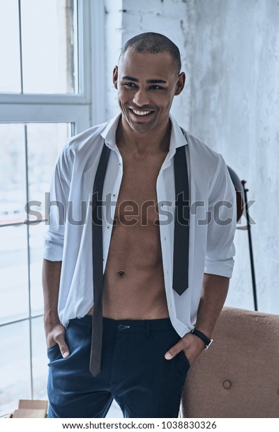 Real\
macho. Handsome young African man in fully unbuttoned white shirt\
looking at camera and smiling while standing at\
home